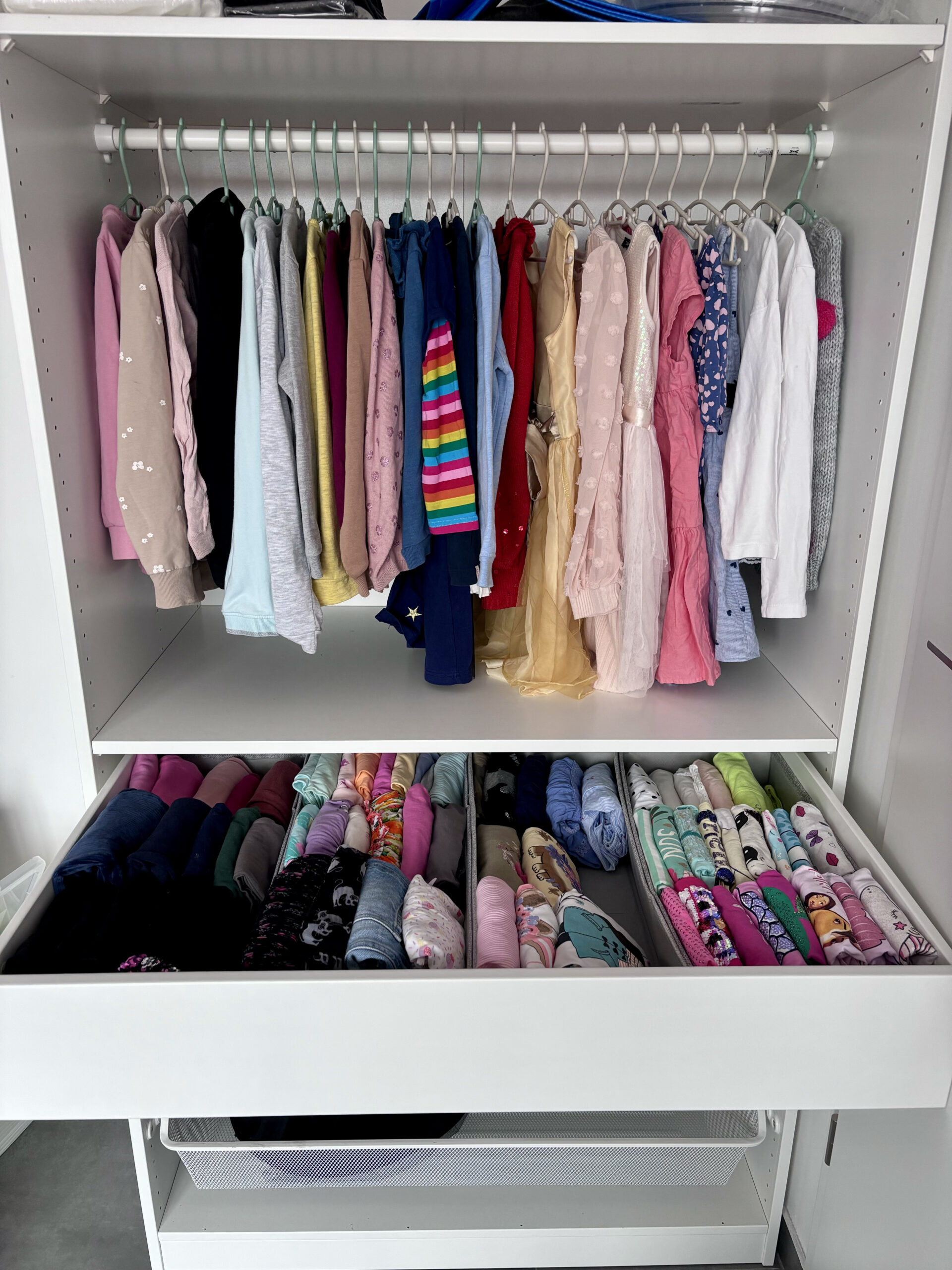 Ein weißer Kleiderschrank, ideal zum Ordnen der Kleidung, mit bunter Kinderkleidung an einer Kleiderstange über einer Schublade mit ordentlich gefalteter Kleidung.