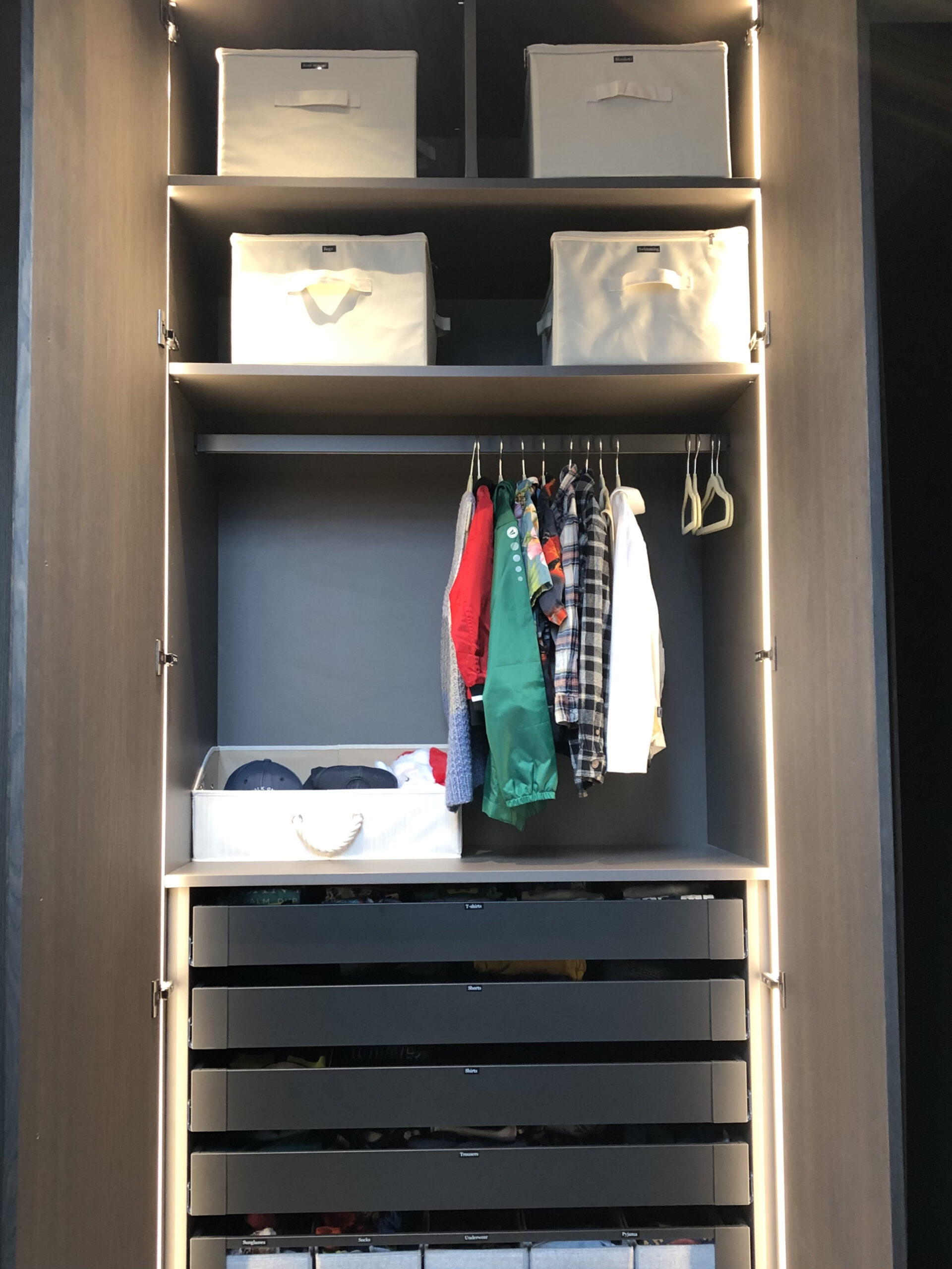 Ein dunkler Schrank, der früher als Kleiderschrank diente, mit Ablagefächern oben, hängender Kleidung, einem Fach für Hüte und vier Schubladen darunter.