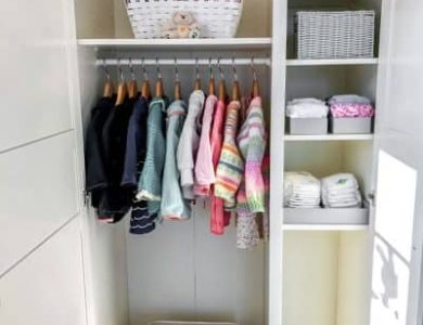 Offener Kleiderschrank mit Kinderkleidung auf Kleiderbügeln, einem Korb darauf und gefalteten Gegenständen auf Regalen. Darin steht ein kleiner Hocker.