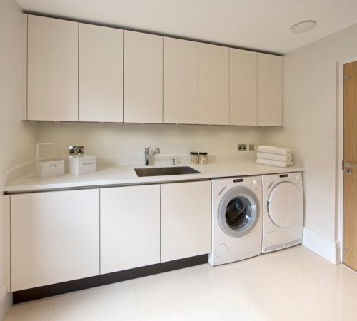 Ein moderner Waschraum mit weißen Schränken, einem Waschbecken sowie übereinander angeordneter Waschmaschine und Trockner zeugt von perfekter Haushaltsorganisation. Rechts befindet sich eine Tür, während ordentlich gefaltete Handtücher die Theke zieren und dem Raum einen Hauch von Ordnung verleihen.